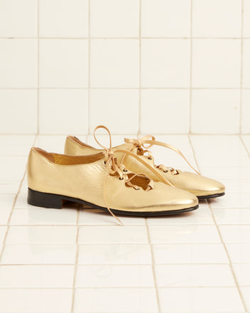 A pair of County Clares - Metallic shoes, crafted from metallic-treated leather with decorative cutouts and tan laces, placed on a white tiled floor.