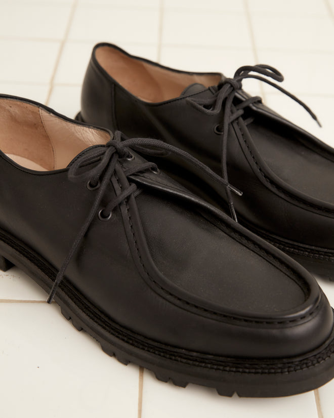 A pair of University Shoes - Black, crafted from Napa leather with a thick rubber sole, is placed side by side on a white tiled floor.