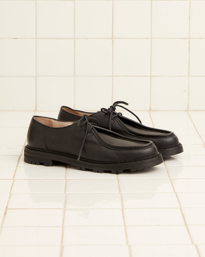 A pair of University Shoes - Black, crafted from Napa leather with a thick rubber sole, is placed side by side on a white tiled floor.