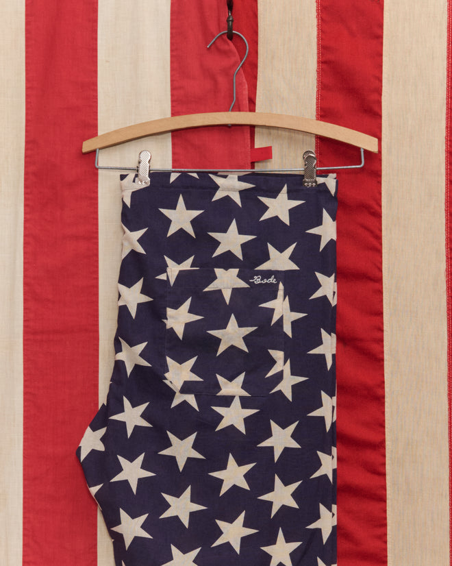 Starry Pajama Pants, crafted from 100% cotton, hang against a red and white striped background. Their blue fabric is decorated with crisp white stars.