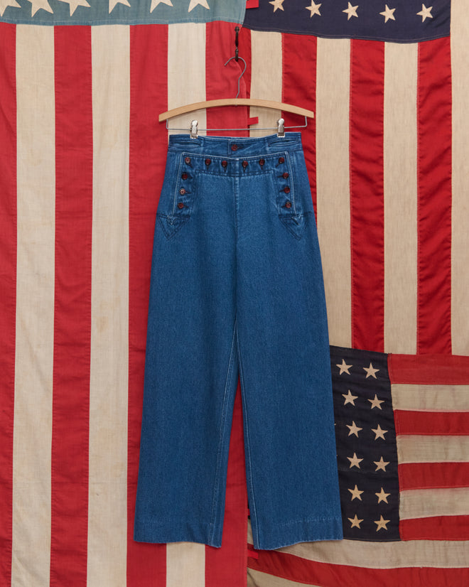 Denim Sailor Trousers in high-waisted blue midweight denim with buttons are displayed on a wooden hanger against an American flag backdrop.