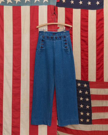 Denim Sailor Trousers in high-waisted blue midweight denim with buttons are displayed on a wooden hanger against an American flag backdrop.