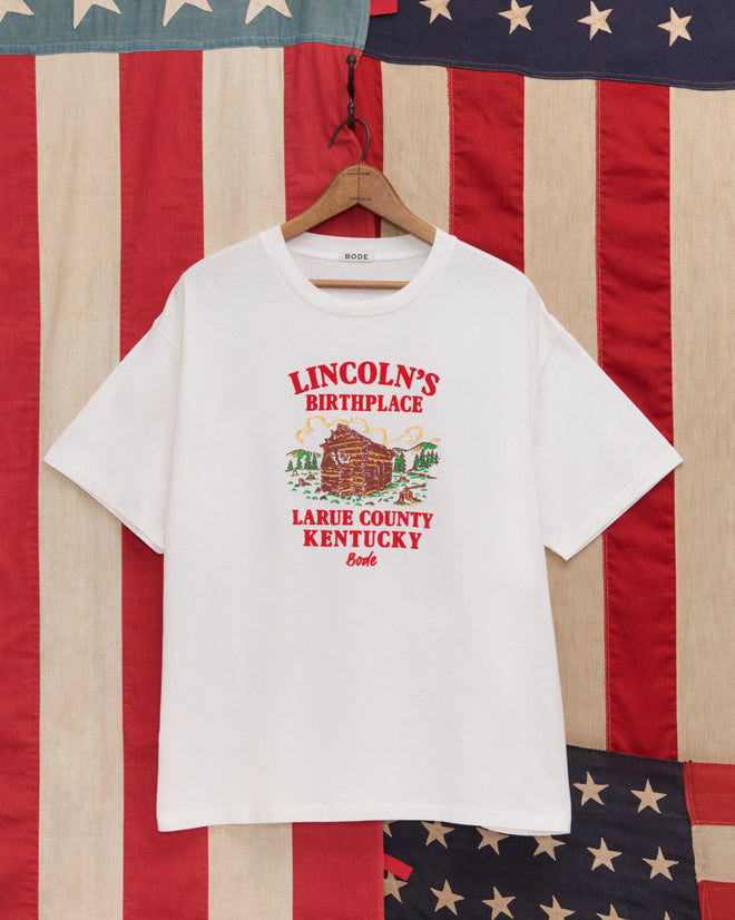 The Lincoln's Birthplace Tee is a white souvenir shirt with “Lincoln’s Birthplace, Larue County, Kentucky” and a log cabin illustration, displayed in front of an American flag.