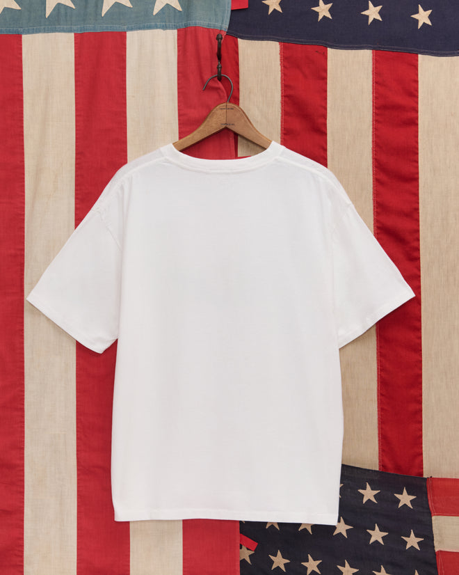 The Lincoln's Birthplace Tee, a plain white souvenir shirt on a hanger, is displayed before an American flag backdrop and features a subtle log cabin illustration inspired by Lincoln’s birthplace.