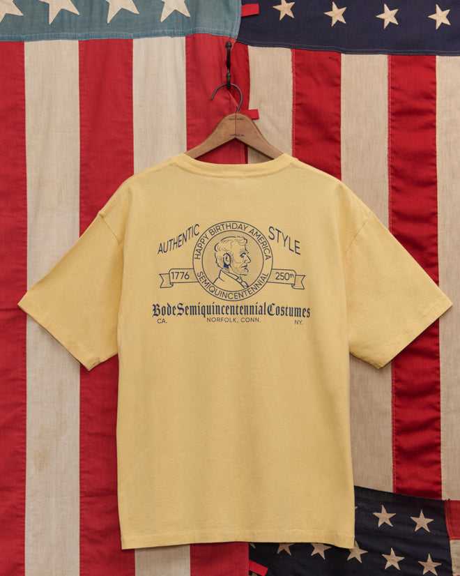 The Lucky Penny Tee, featuring a vintage-style yellow Lincoln penny graphic, hangs on a wooden hanger in front of an American flag, showcasing authentic style.