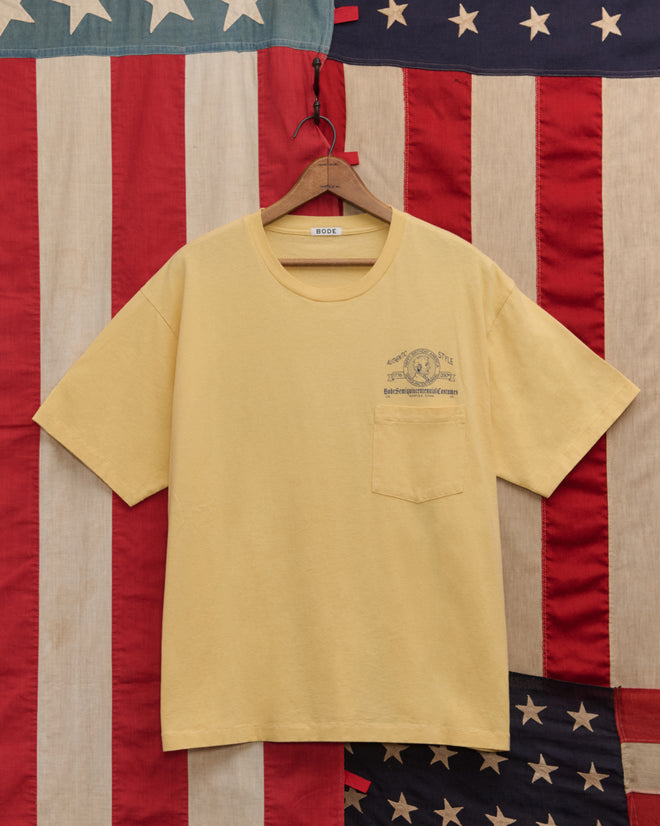 A yellow Lucky Penny Tee with a chest pocket hangs in front of a vintage American flag backdrop, capturing authentic style and timeless Americana.