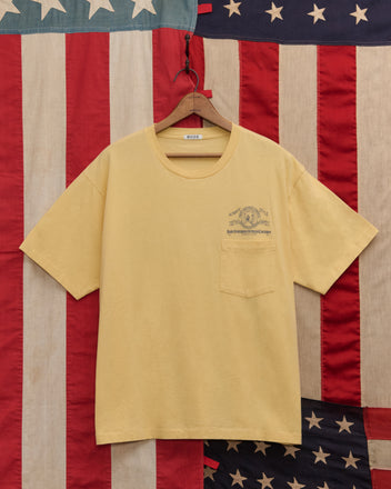 A yellow Lucky Penny Tee with a chest pocket hangs in front of a vintage American flag backdrop, capturing authentic style and timeless Americana.