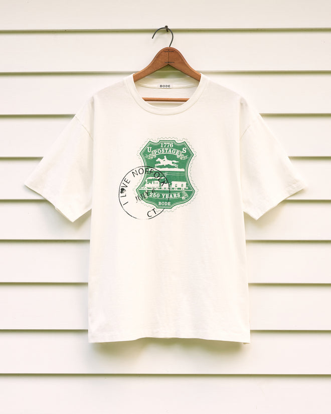 A white “Norfolk Stamp Tee” on a hanger featuring a green “1776 Utopiats” graphic and partial black “I LOVE NO” text, reminiscent of an American centennial tee.