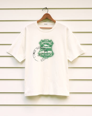 A white “Norfolk Stamp Tee” on a hanger featuring a green “1776 Utopiats” graphic and partial black “I LOVE NO” text, reminiscent of an American centennial tee.