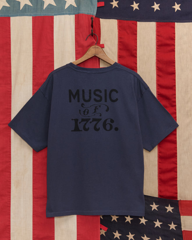 The Lincoln Lantern Tee, featuring "MUSIC OF 1776." text, hangs on a wooden hanger against an American flag background, capturing the spirit of torchlight parades and historic celebrations.
