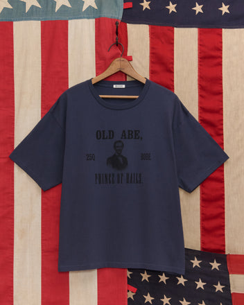 The Lincoln Lantern Tee, a navy blue T-shirt with "OLD ABE, PRINCE OF RAILS," hangs before an American flag backdrop, recalling Abraham Lincoln’s historic campaign parades.