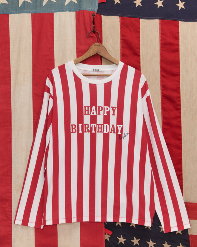 The Birthday Stripe Tee features red and white stripes with "HAPPY BIRTHDAY" text, displayed in front of an American flag—ideal for celebrating America’s 250th birthday.