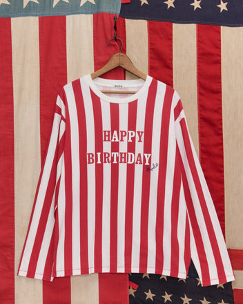 The Birthday Stripe Tee features red and white stripes with "HAPPY BIRTHDAY" text, displayed in front of an American flag—ideal for celebrating America’s 250th birthday.