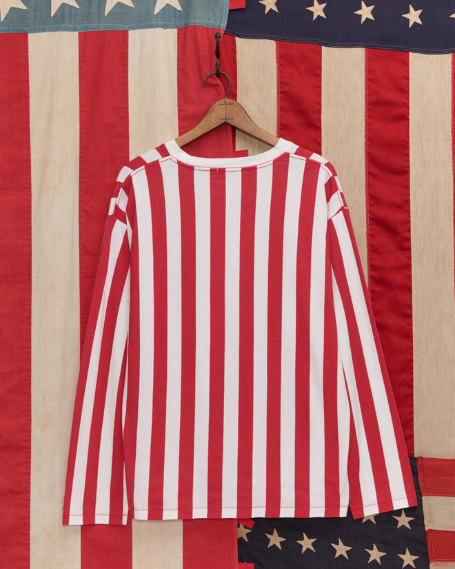 The Birthday Stripe Tee, featuring red and white vertical stripes, hangs before an American flag backdrop—perfect for celebrating America’s 250th birthday in style.
