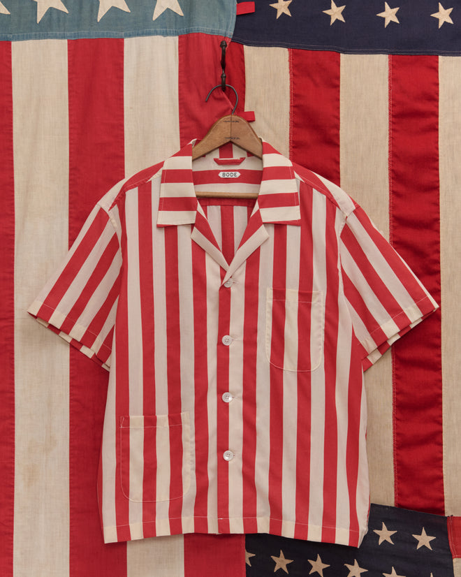 The Valance Stripe Short Sleeve Shirt, featuring red and white stripes and made from 100% cotton, hangs against an American flag backdrop, offering a vintage-inspired look that combines classic style with comfort.