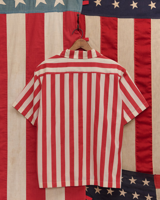 The Valance Stripe Short Sleeve Shirt, crafted from 100% cotton with vintage-inspired red and white stripes, hangs before American flag fabrics, evoking timeless Americana.
