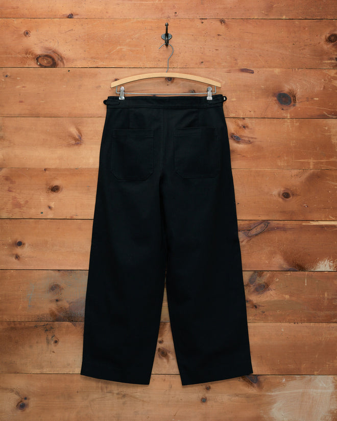 The Wide Leg Snap Trousers - Black are crafted from 100% cotton heavyweight twill and evoke the classic look of vintage Australian military pants. These high-waisted trousers feature back pockets and are elegantly displayed on a wooden hanger against a wood wall background.