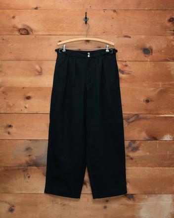 Wide Leg Snap Trousers - Black, high-waisted and heavyweight, are crafted from 100% cotton twill and hang on a hanger against a rustic wooden wall—evoking the timeless appeal of vintage Australian military pants.