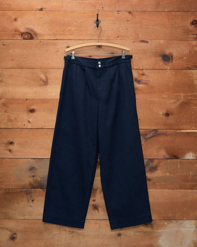 Wide Leg Snap Trousers - Navy displayed on a wooden hanger against a rustic wooden wall background.