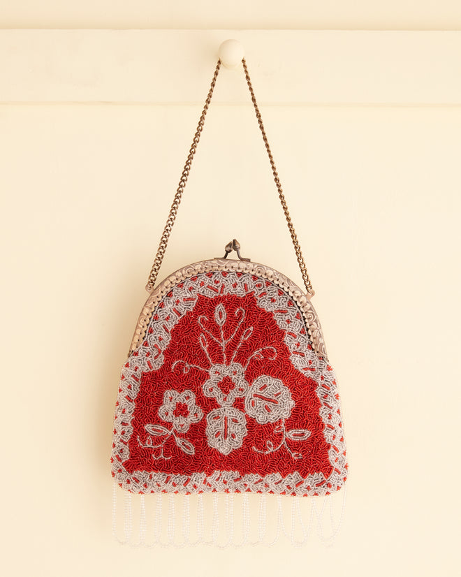 The Amrita Beaded Bag, featuring a vintage-style red and white floral pattern, hangs on a white wall by a metal chain.