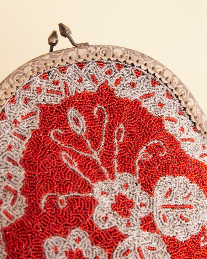 The Amrita Beaded Bag, featuring a vintage-style red and white floral pattern, hangs on a white wall by a metal chain.