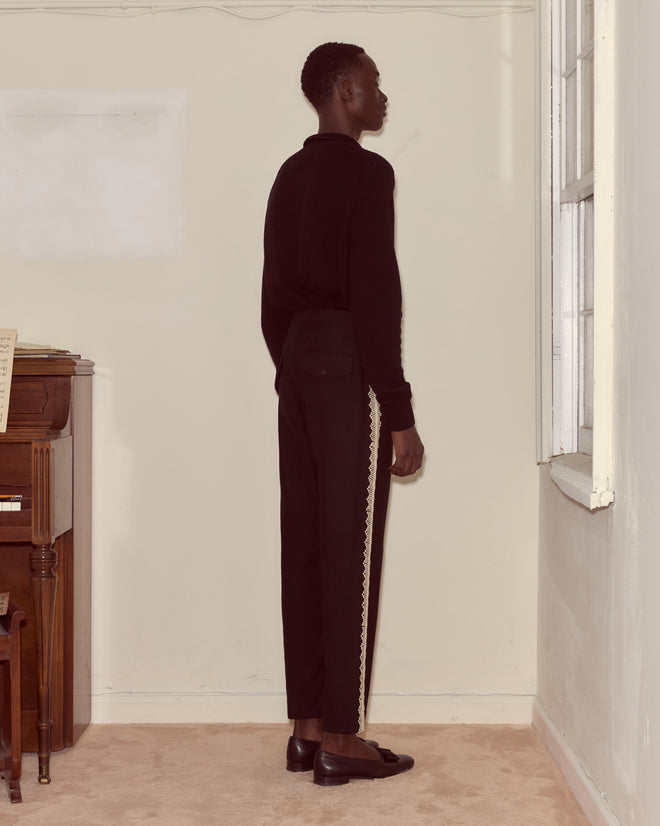 A man wearing Lacework Side-Buckle Trousers stands sideways, facing a window in a simple beige room with a piano.