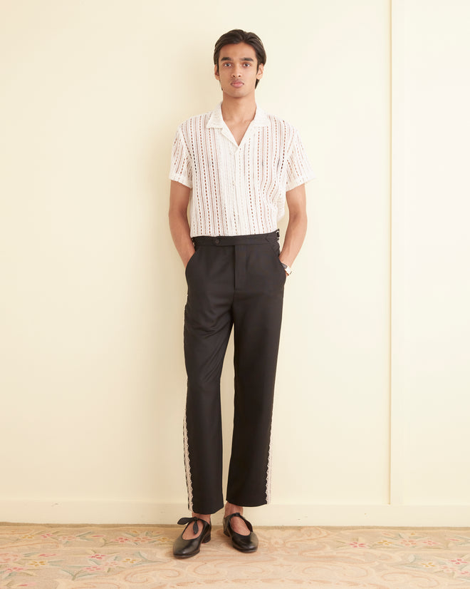 A person stands against a beige wall wearing a white short-sleeve shirt, black Lacework Side Buckle Trousers with side-buckle waist adjusters, and black shoes with their hands in their pockets.