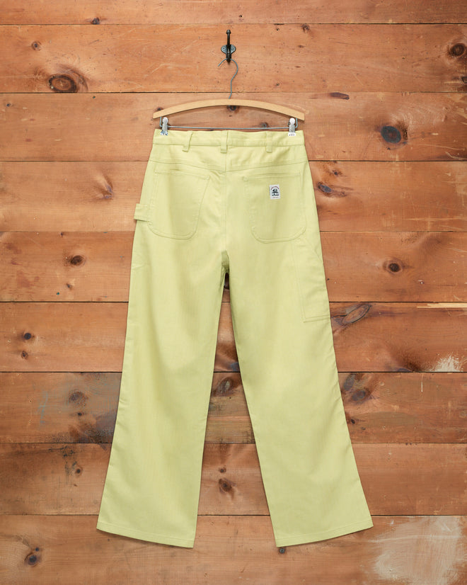 The Herringbone Knolly Brook Trousers in green, featuring front pockets, are displayed on a wooden hanger against a rustic wooden wall background.