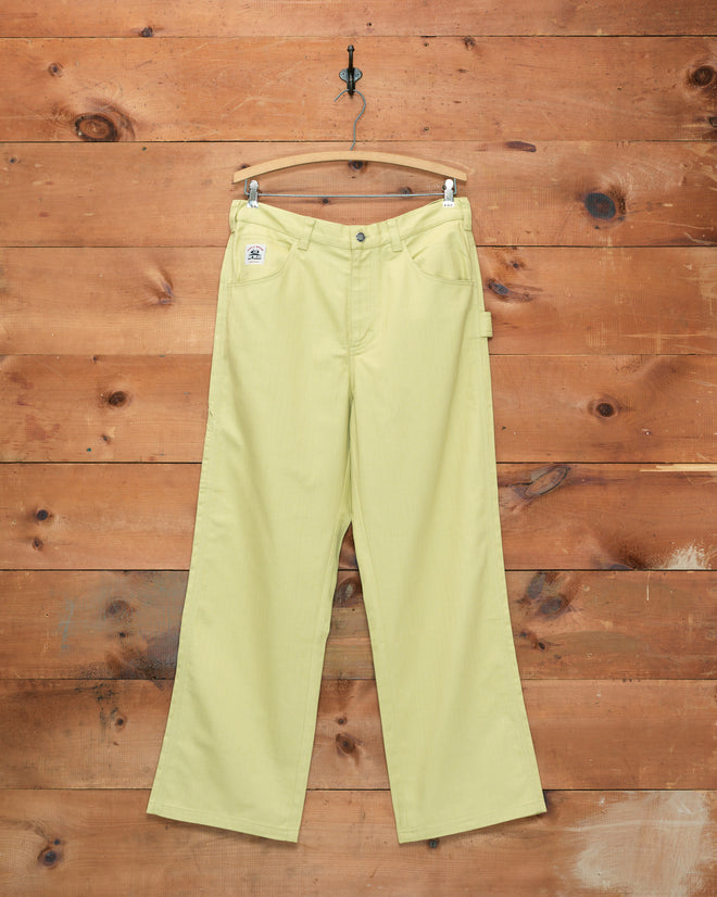 The Herringbone Knolly Brook Trousers in green, featuring a wide-leg silhouette, hang on a wooden hanger against a rustic wooden wall.