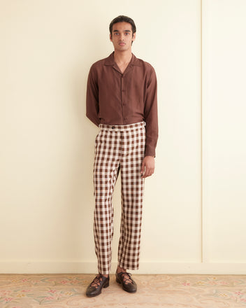 A person stands indoors against a plain wall, wearing a brown button-up shirt and the Gilbert Check Trousers, which feature 1950s-inspired checkered patterns and side-buckle waist adjusters. They have their hands in their pockets and are sporting dark lace-up shoes.