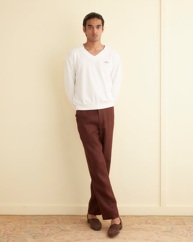 A person wearing a white Boston Terry Pullover and brown pants, which appear to be from the embroidered Bode collection, stands against a cream-colored wall with arms behind their back. They are standing on a beige floor with a subtle floral pattern.