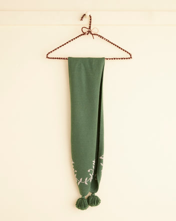 The Floret Scarf in green, featuring embroidered white floral patterns and tassels, gracefully hung on a twisted wire hanger against a light wall.