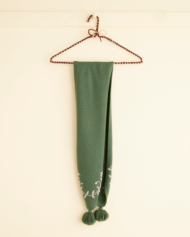 The Floret Scarf in green, featuring embroidered white floral patterns and tassels, gracefully hung on a twisted wire hanger against a light wall.