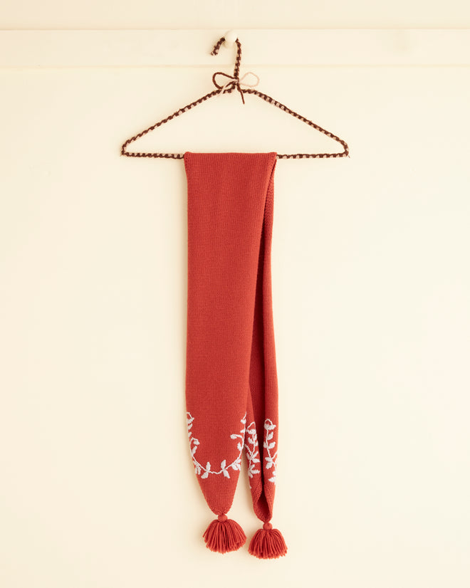 The Floret Scarf - Red, featuring embroidered floral designs and tassels at both ends, is displayed on a braided hanger against a beige wall.