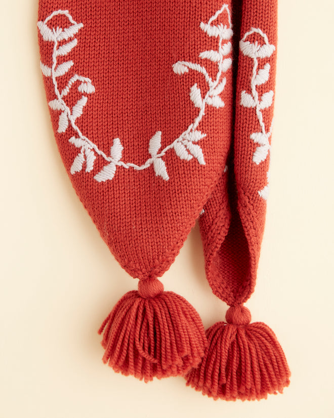 The Floret Scarf - Red, featuring embroidered floral designs and tassels at both ends, is displayed on a braided hanger against a beige wall.