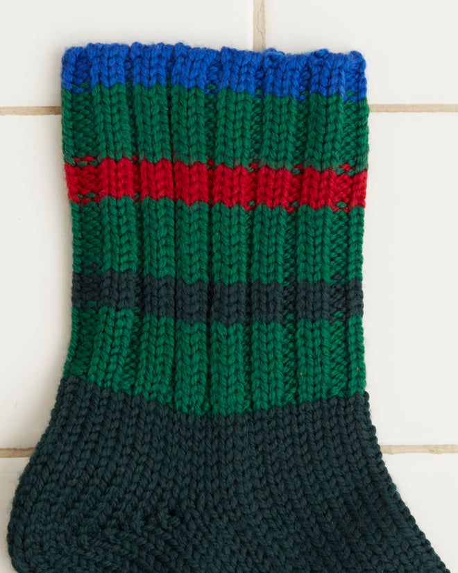 The meticulously hand-knit Herrington Stripe Socks in dark green, crafted from merino wool, boast cozy red and green striped cuffs and are showcased against a white tiled background.