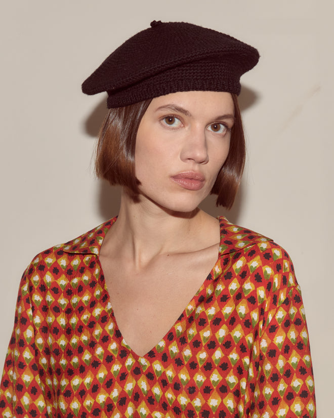 A woman with short brown hair looks forward, wearing a black Crochet Beret and a patterned red, yellow, and black top.