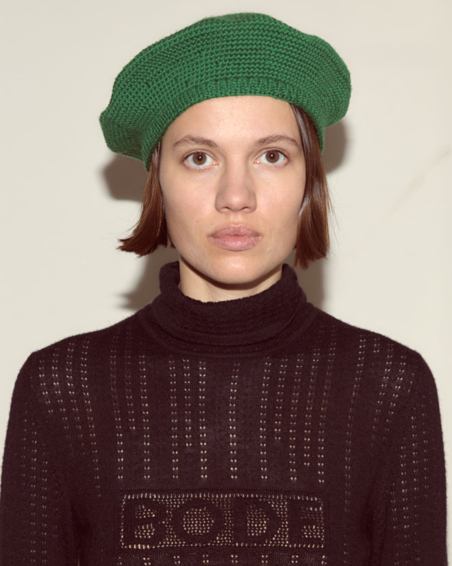 Person with short brown hair wears a green Crochet Beret and black sweater, facing forward against a plain background.