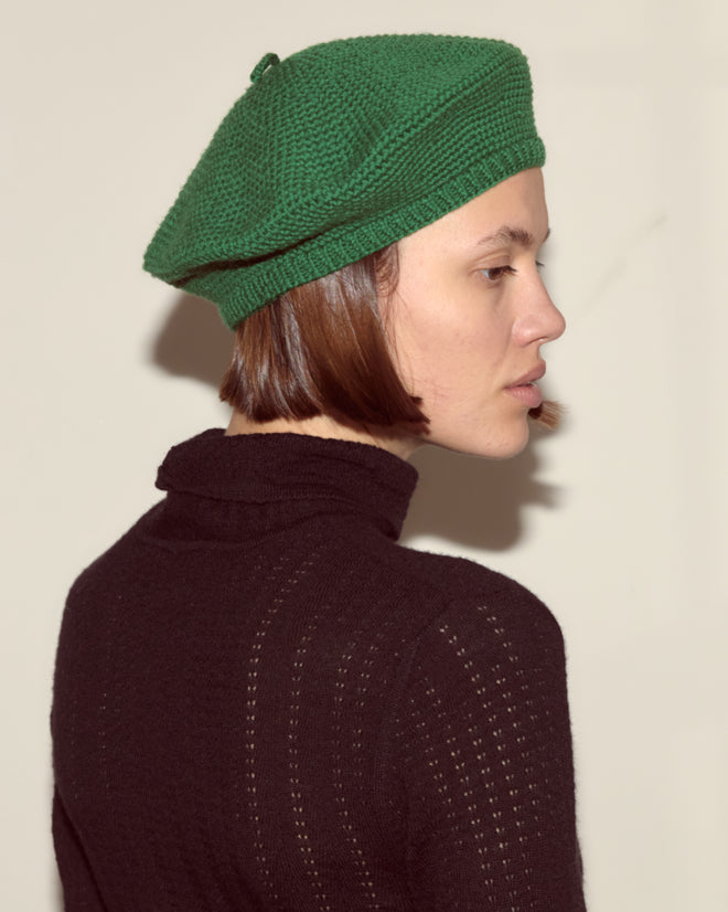 A person in a green Crochet Beret and black turtleneck sweater faces sideways against a plain background, highlighting timeless accessories.