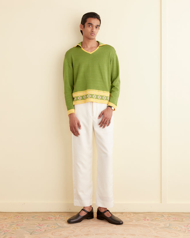 A person stands against a light-colored wall, wearing the Wellfleet Jacquard Polo, a green sweater with yellow details knit from fine cotton yarn, paired with white trousers and dark shoes. Their left hand is relaxed by their side.