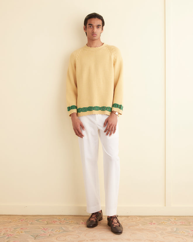 A person stands against a cream-colored wall, wearing the Daisy Garland Sweater in yellow, paired with white pants and brown shoes.