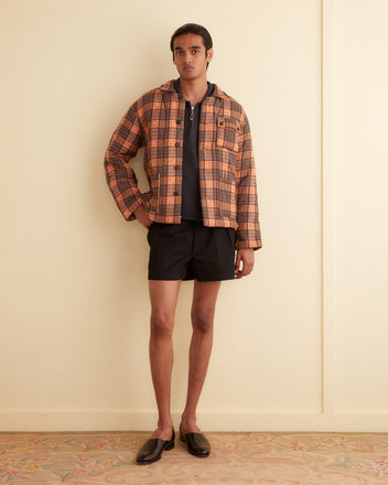 A person stands indoors wearing a Russet Plaid Quilt Jacket crafted from 1930s-1950s deadstock plaid fabric. They pair it with a dark shirt, black shorts, and black shoes. With one hand in their pocket, they look directly at the camera.