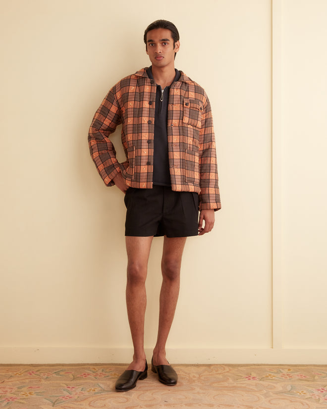 A person stands indoors wearing a Russet Plaid Quilt Jacket crafted from 1930s-1950s deadstock plaid fabric. They pair it with a dark shirt, black shorts, and black shoes. With one hand in their pocket, they look directly at the camera.