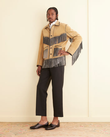 A person stands against a wall, wearing the Duo Suede Appalachian Jacket adorned with hand-cut fringe details, complemented by a white shirt, black pants, and black slip-on shoes.