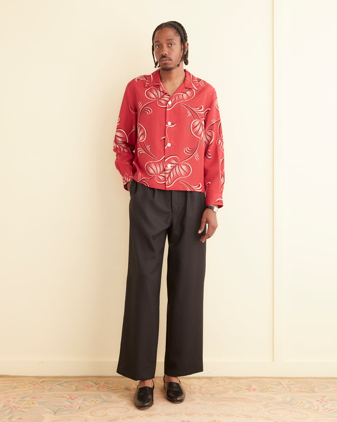 Person standing indoors wearing the Creeping Begonia Long Sleeve Shirt, which is red with a botanical print and made from 100% viscose in Portugal. They are also wearing black wide-leg trousers and black shoes. Light-colored walls and a patterned rug are in the background.