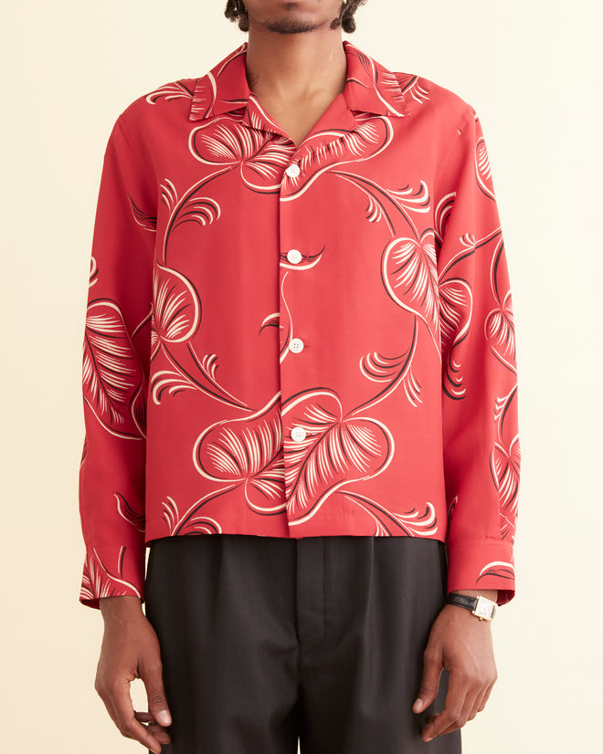 Person standing indoors wearing the Creeping Begonia Long Sleeve Shirt, which is red with a botanical print and made from 100% viscose in Portugal. They are also wearing black wide-leg trousers and black shoes. Light-colored walls and a patterned rug are in the background.