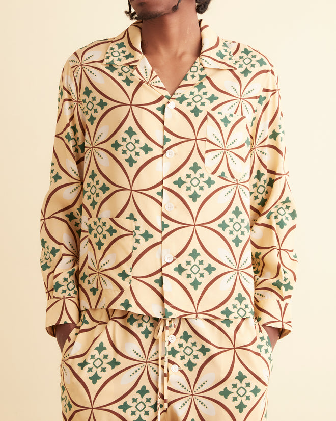 A man stands against a plain wall, wearing the Greer Long Sleeve Shirt paired with matching bottoms. The set features a 1960s style geometric pattern in beige, green, and orange tones. He is looking directly at the camera with a neutral expression.