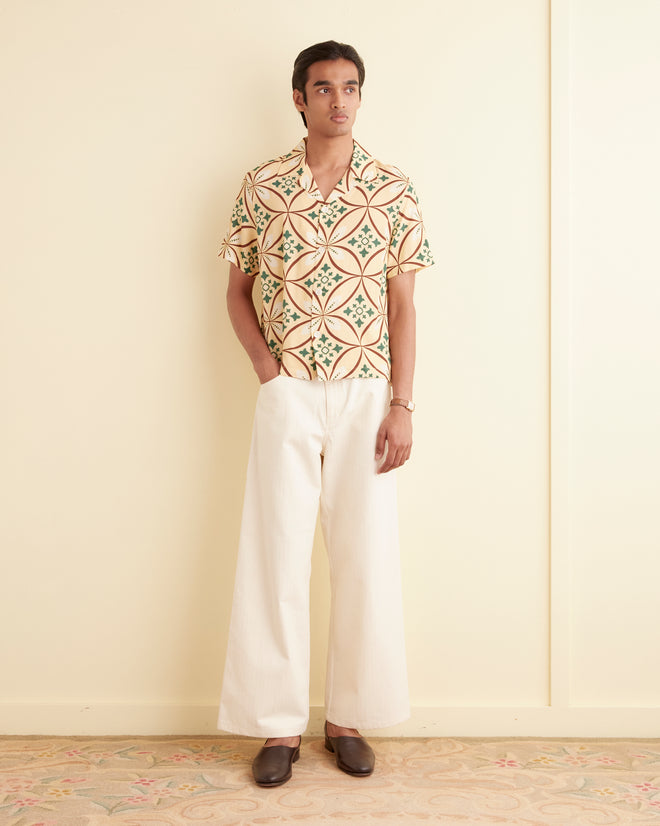 A man stands against a cream-colored wall, wearing the Greer Short Sleeve Shirt with a geometric pattern from the 1960s, white wide-leg pants, and brown shoes. His hands are in his pockets.