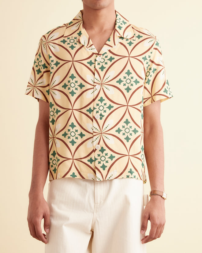 A man stands against a cream-colored wall, wearing the Greer Short Sleeve Shirt with a geometric pattern from the 1960s, white wide-leg pants, and brown shoes. His hands are in his pockets.