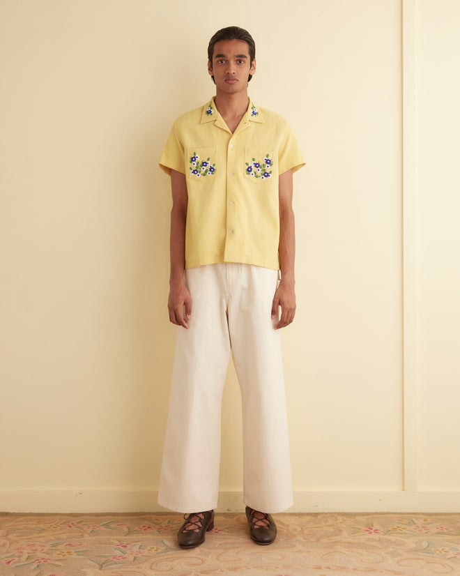 A person stands against a plain wall, wearing a Beaded Chicory Short Sleeve Shirt in yellow, adorned with floral beading, paired with 100% cotton white wide-leg pants. They have long hair and brown shoes. The floor features a floral patterned rug.